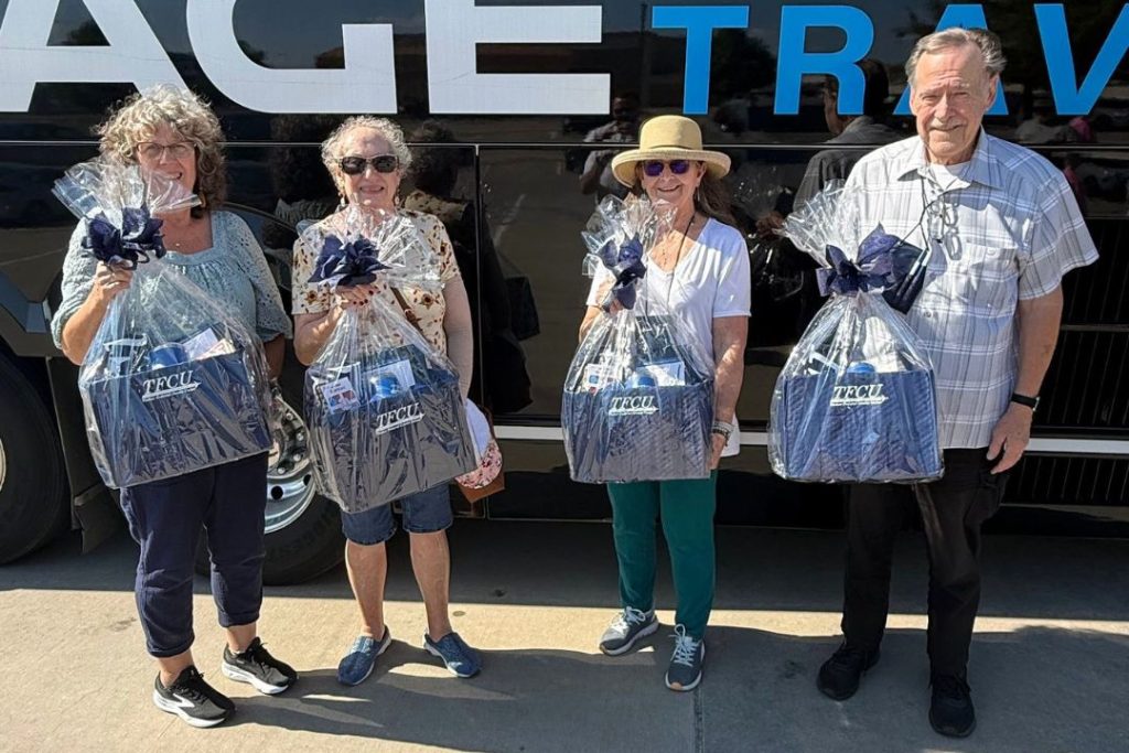 Four older people hold TFCU gift baskets in front of a bus