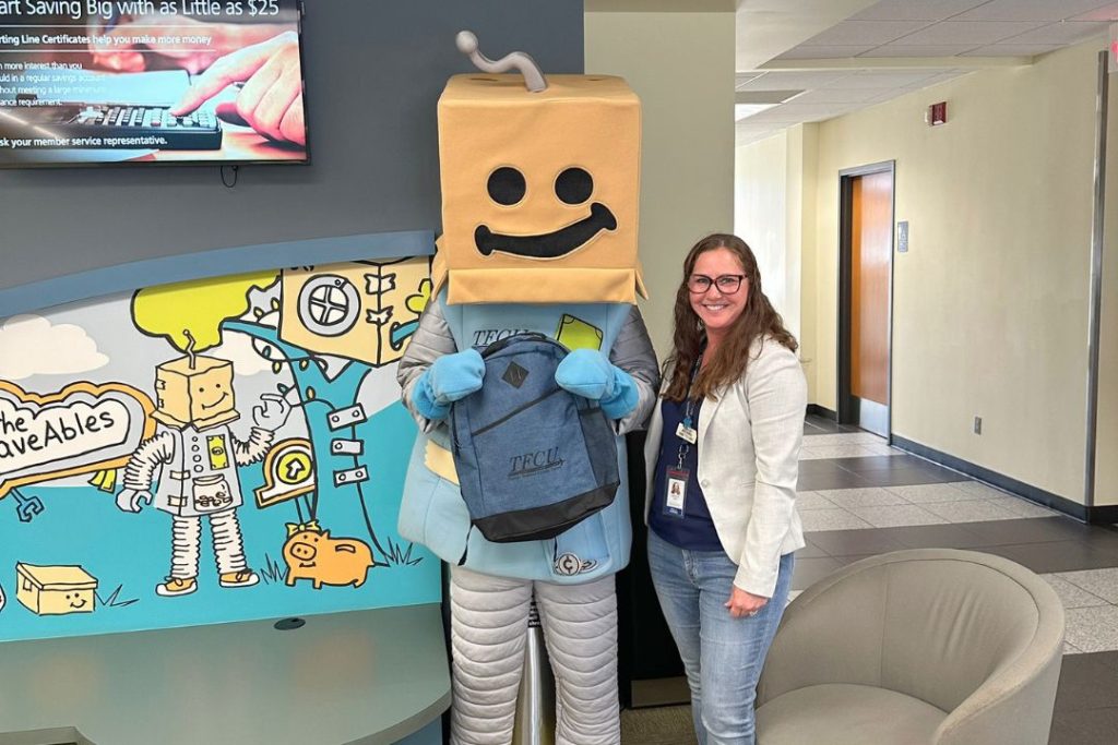 SaveATron and a TFCU employee stand in a TFCU branch holding a backpack