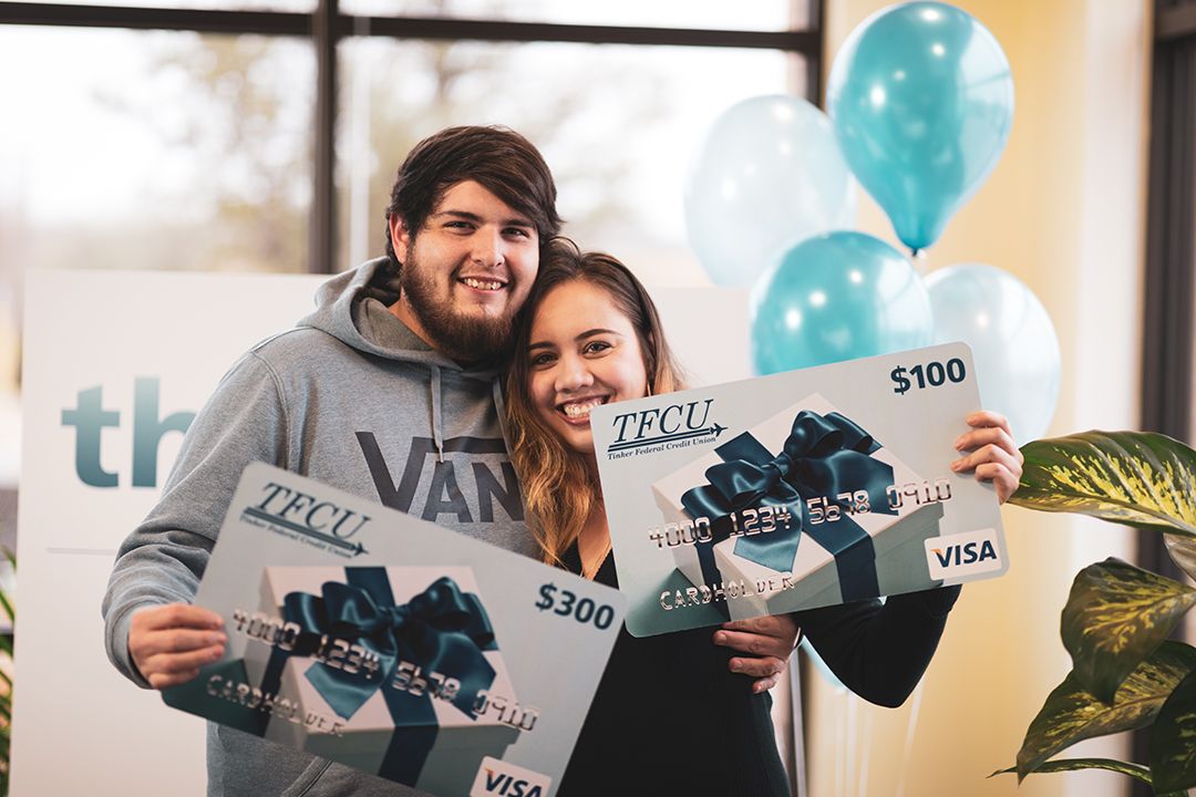 A couple who are TFCU members are both holding a TFCU gift card sign, hugging, and smiling.