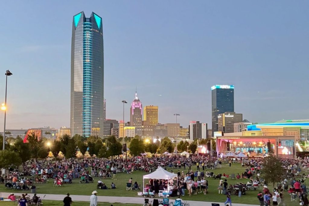A crowded park with a music stage overlooking the Oklahoma City skyline