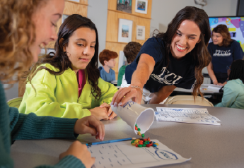 Students seated at a classroom table working on worksheets while an instructor pours colorful counting pieces from a cup for a hands-on financial education activity.