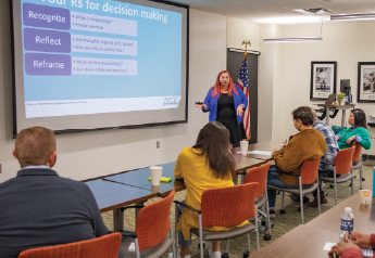 Instructor presenting at the front of a classroom using a projected slide while attendees sit at tables and take notes during a financial education workshop.