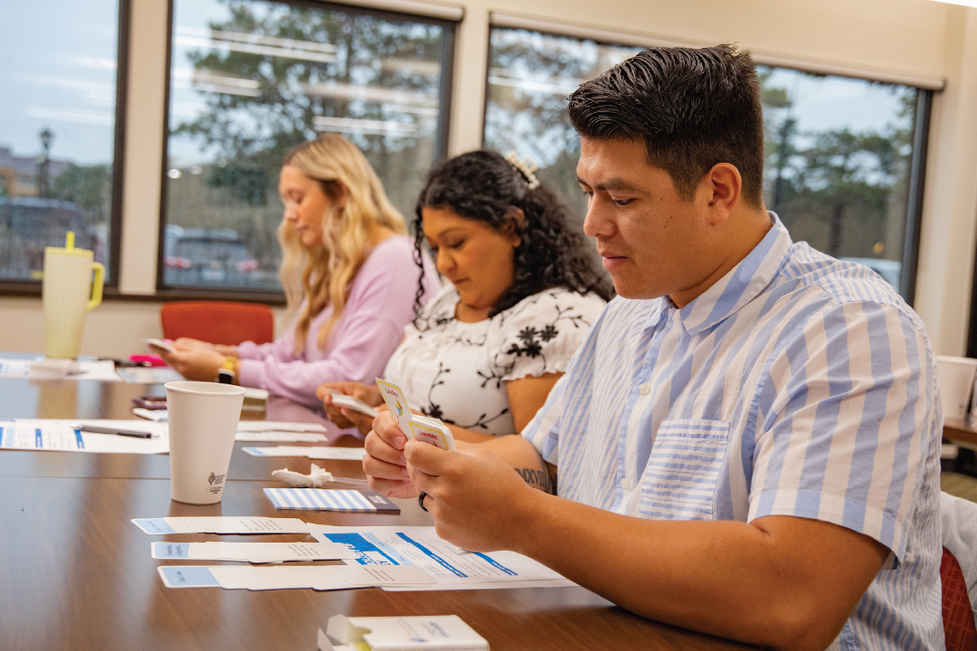 Three people sit at a desk and look at flash cards and worksheets