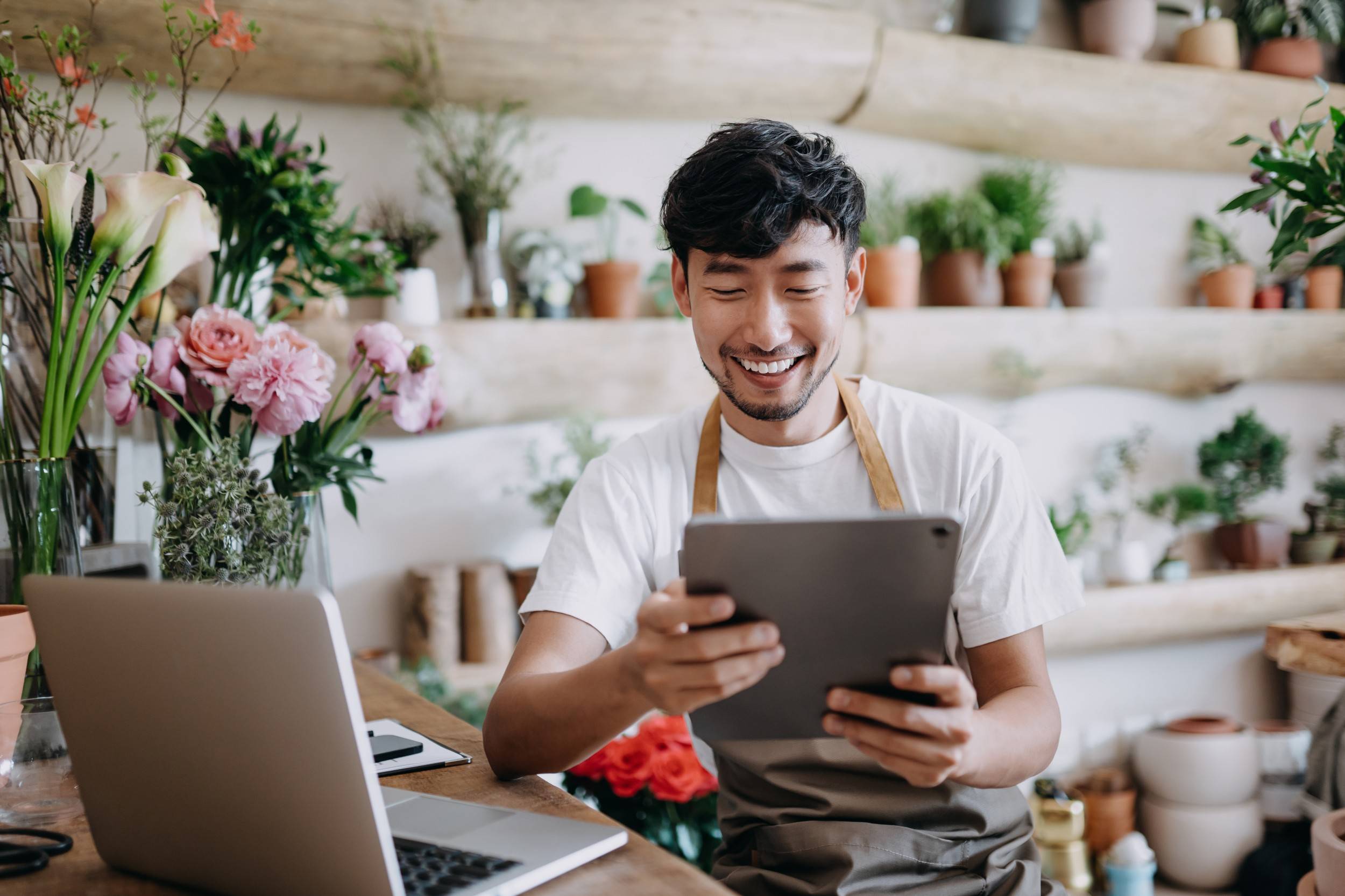 A smiling shop owner looks at his tablet with a laptop nearby sitting on the counter.