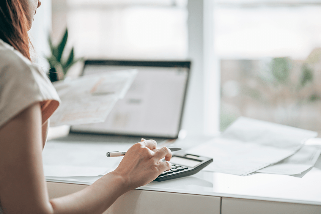 Close up of someone typing on a calculator with a laptop in the back