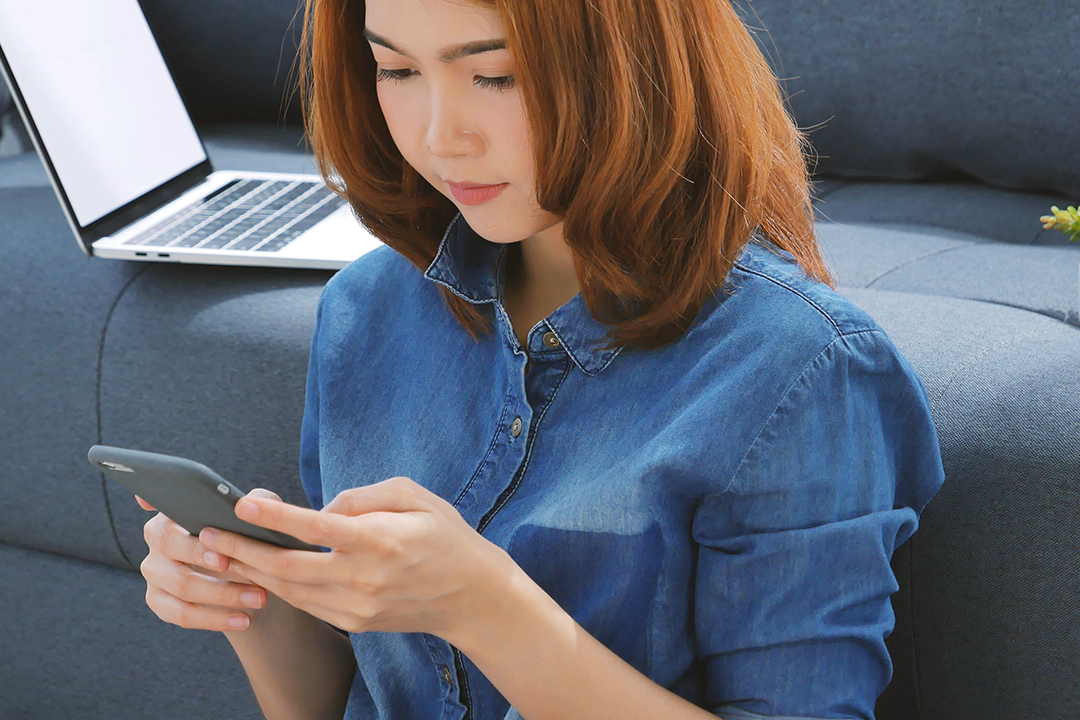 Young woman sitting on the floor on her smartphone with laptop in background
