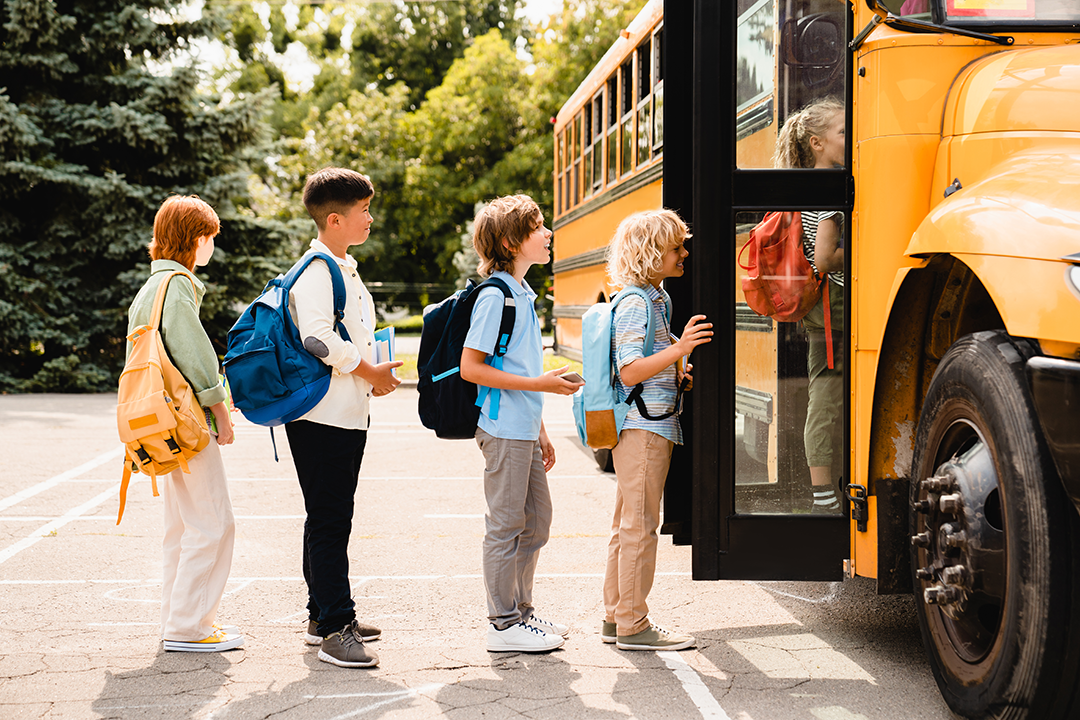 Four students wearing backpacks lining up to get onto a school bus.