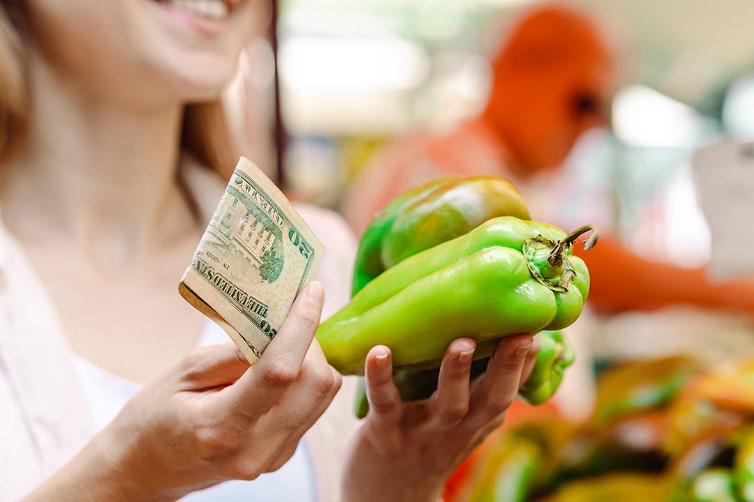 Close up of a woman holding a $20 bill and a handful of green bell peppers