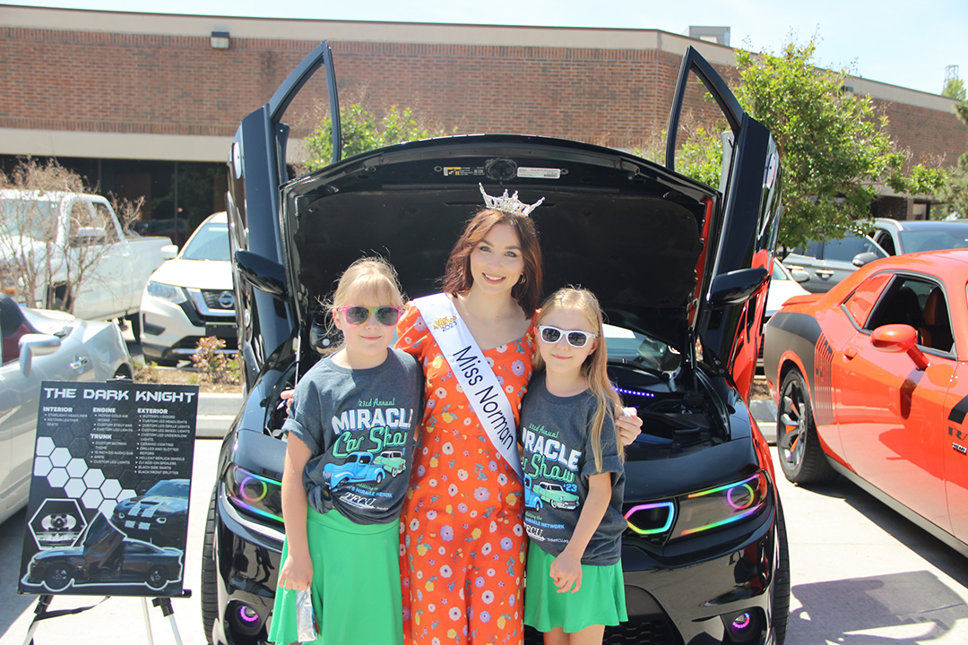 Miss Norman, Sunny Day, wearing a crown and sash, poses with Miracle Children, Vivianne and Veronica in front of a shiny black sports car with doors raised vertically and neon lights glowing.