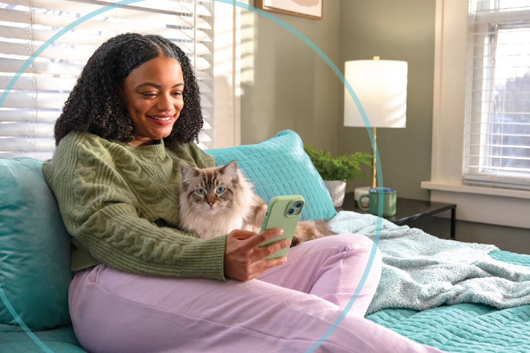 A young girl on her mobile device sitting on a bed with her cat.