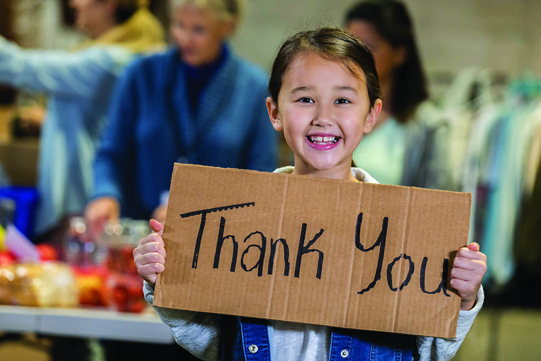 Young girl holding a cardboard sign that says "thank you" and smiling with people in the background.