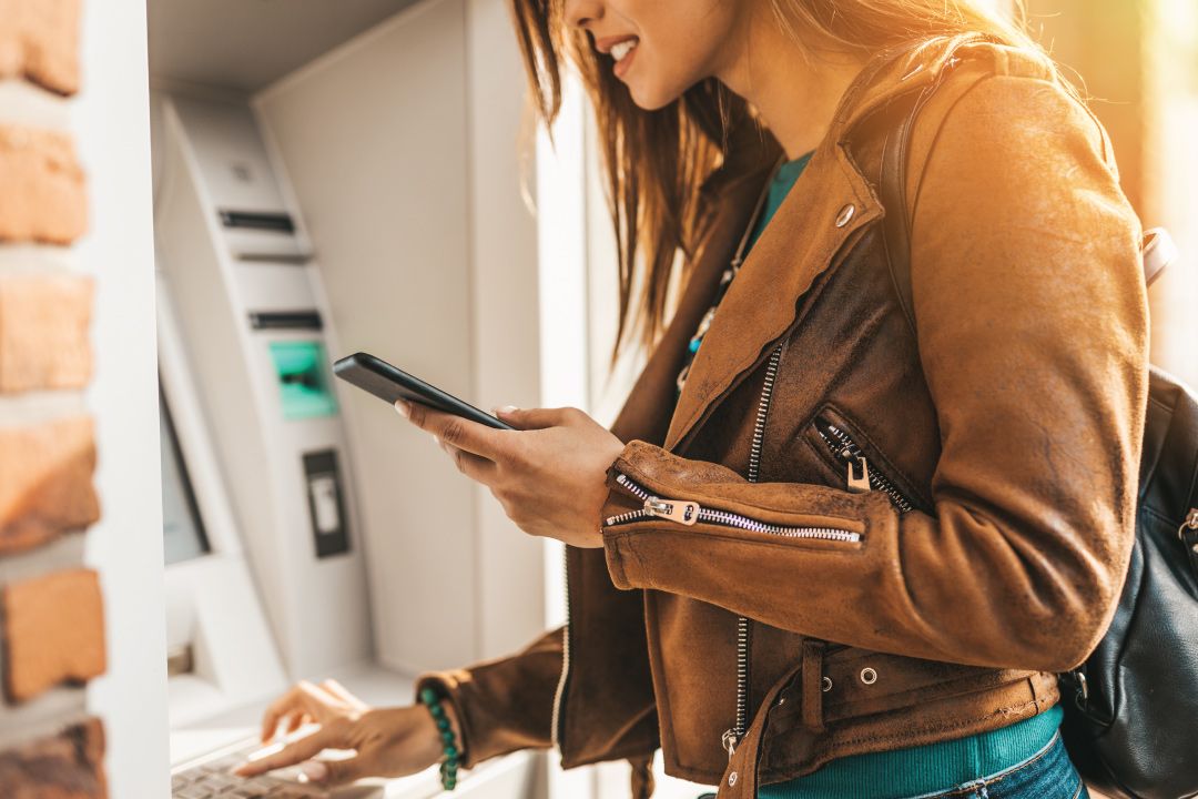 Young Female at atm machine withdrawing cash and using smartphone