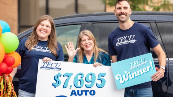 A woman stands with two TFCU staff members holding a sign that says "$7,696 Auto Loan Payoff"