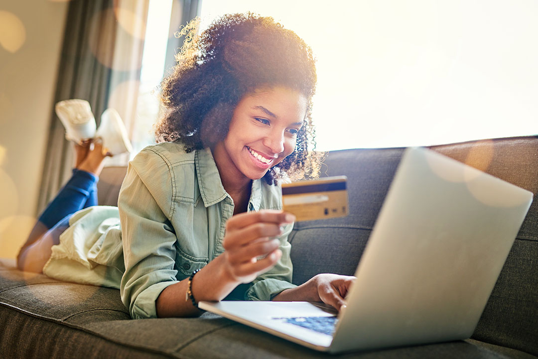 A young women lying on the couch at home while on her laptop and holding a card.