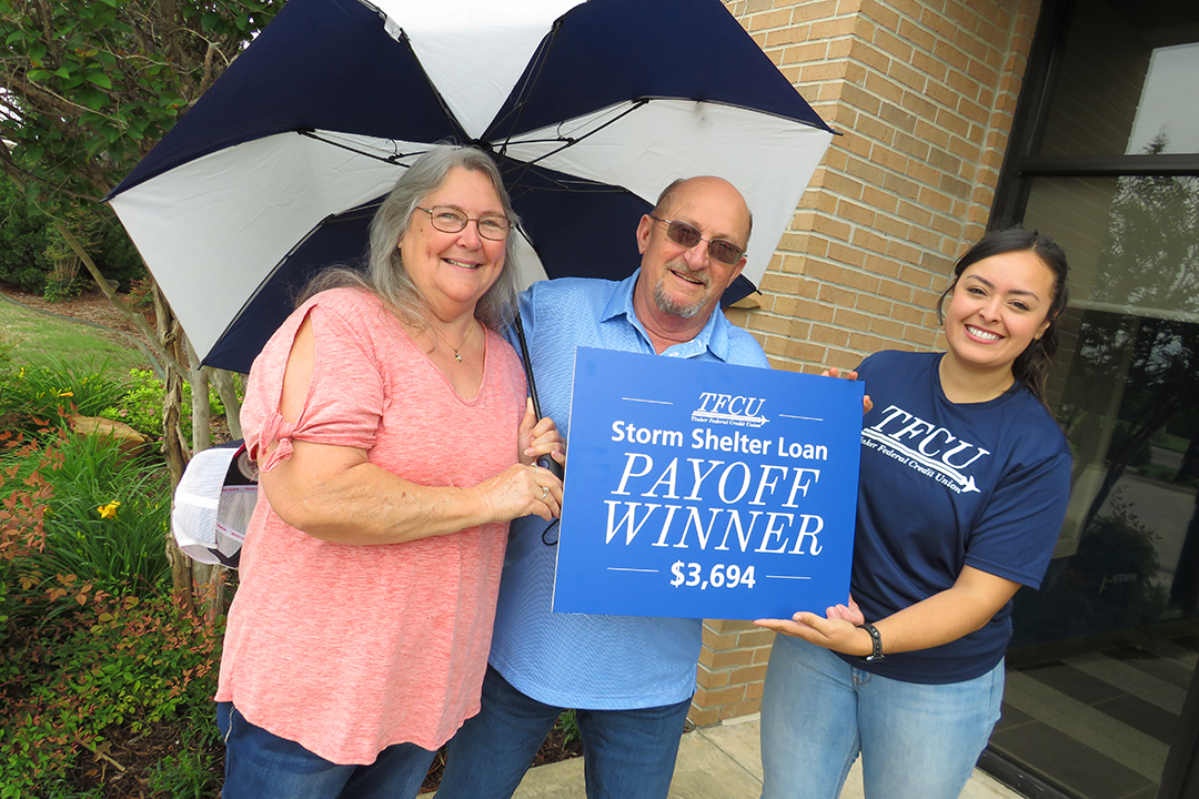 Older man and woman holding a Storm Shelter Payoff Winner sign and umbrella with a TFCU employee.