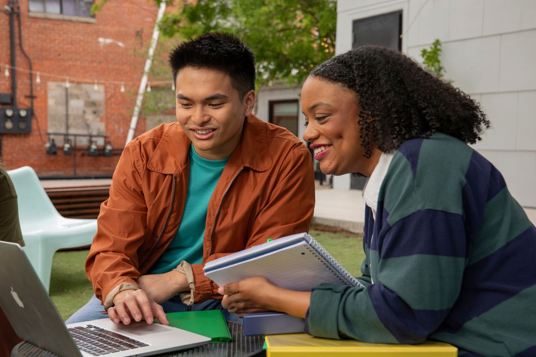 Two college students sit outside with notebooks looking at a laptop.