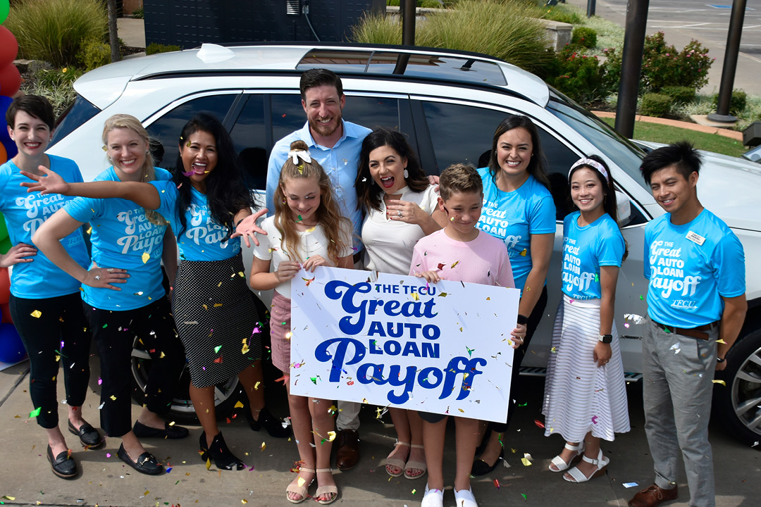 A family with a mom, dad and two younger kids standing with TFCU employees in front of a car and holding a TFCU Great Auto Loan payoff sign.