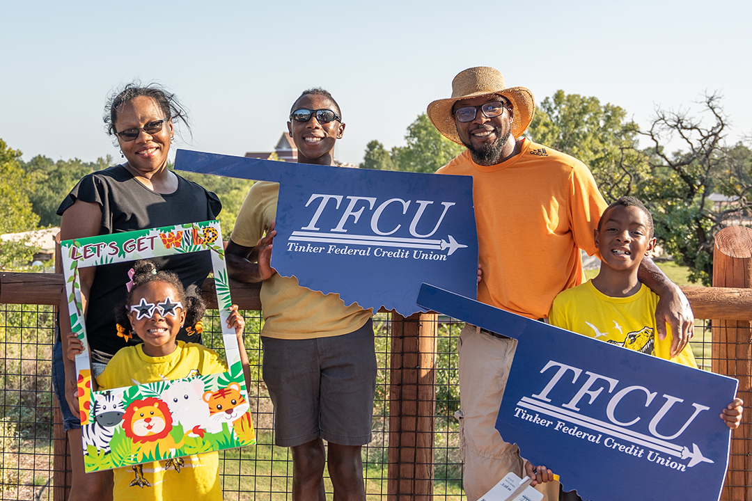 A mom, dad and three kids holding up oklahoma-shaped signs and a zoo photo sign.