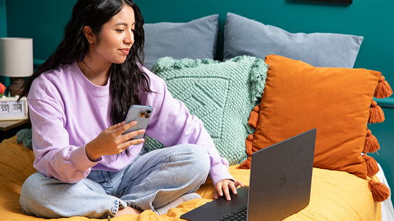A young woman sitting on her bed while on her mobile device and laptops.