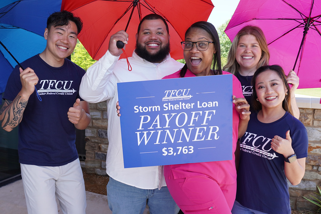 Three TFCU employees standing with a couple while holding umbrellas and a sign that says storm shelter payoff winner