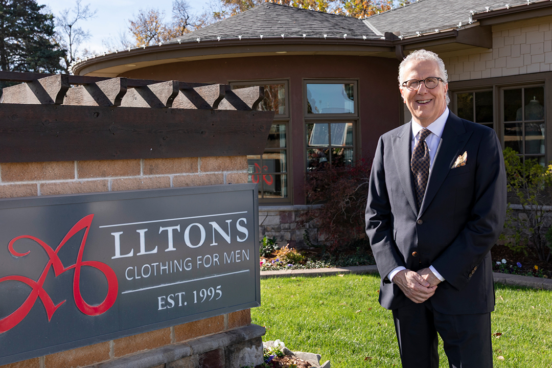 A man in a suite stands beside a sign that says Alltons Clothing for Men