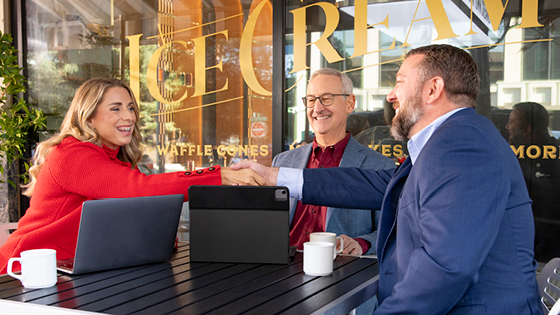 A woman and businessman shake hands while sitting a table outside with another businessman