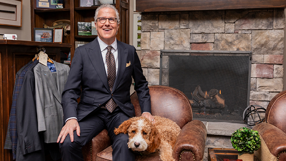 A man sits on the arm of a leather chair with his dog in front of a fireplace