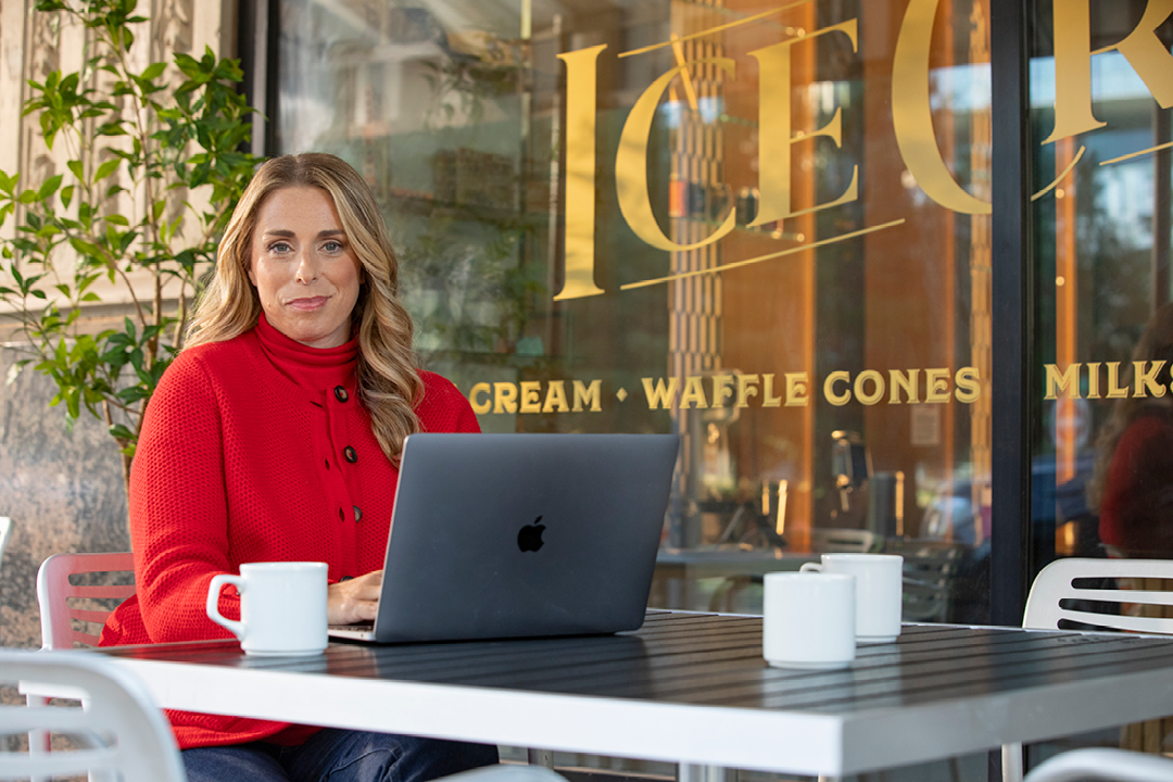 A woman sits outside at a table with her laptop and a cup of coffee