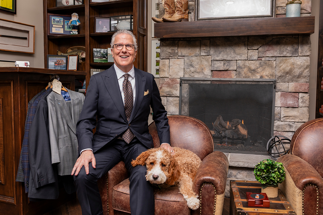 A man in a suit sits on a leather chair in front of a fireplace with his dog
