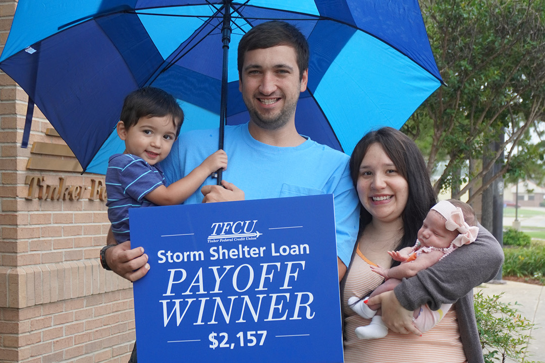 Family holding a blue TFCU sign reading “Storm Shelter Loan Payoff Winner $2,157,” standing under a blue umbrella outdoors.