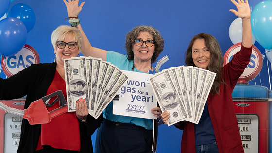 Three women stand together. Two hold large dollar bill cutouts and one holds a sign that says, "I won gas for a year",