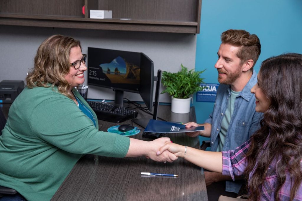 A TFCU employee shakes hands with a man and a woman while sitting at a desk