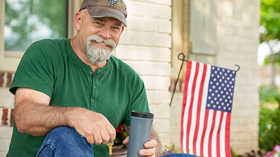 Older veteran sitting in front of his house holding a key with an american flag in the back.