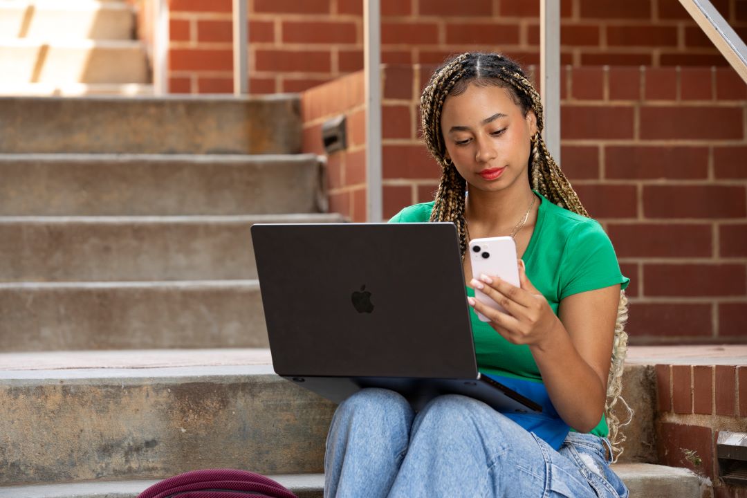 TFCU 72424 2817 A young woman sits on steps outside while looking at her phone and laptop.