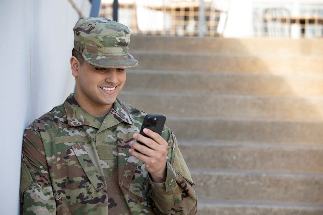 Military service member in uniform using a smartphone while seated beside outdoor steps.