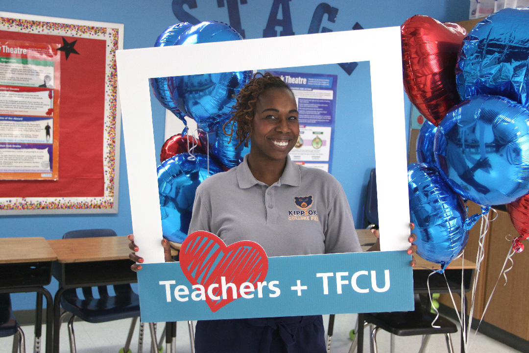 A woman who is a teacher is holding up a sign that says Teachers + TFCU