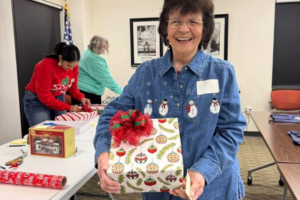 An older woman smiles at the camera while holding a wrapped Christmas gift while two other people are wrapping gifts at a table in the background.