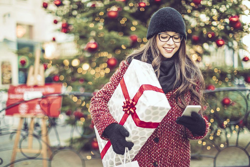 Young woman holding a gift box and shopping bags while on her smartphone.