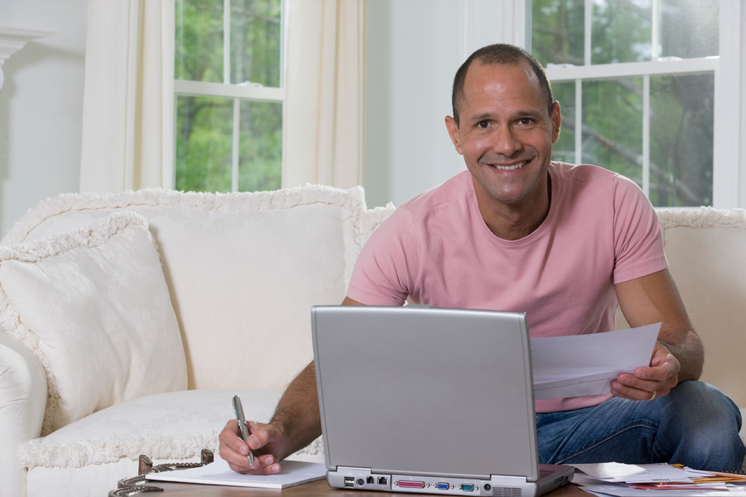 Man in pink t-shirt sitting on white couch and sorting out bills in front of a laptop