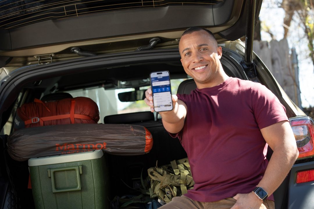 A man holding up a mobile phone while sitting in the trunk of his vehicle