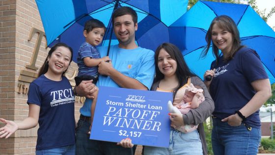 Group of people standing outside under large blue umbrellas, holding a TFCU sign that reads “Storm Shelter Loan Payoff Winner $2,157,” in front of a brick building.