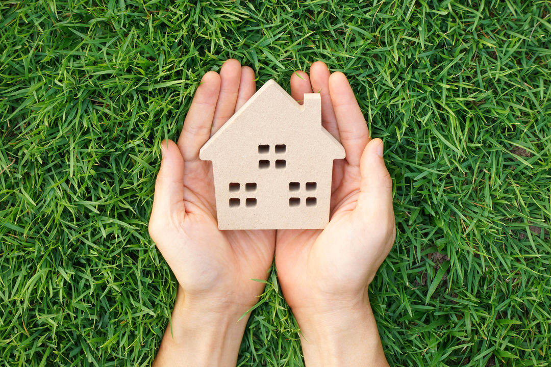 Cupped hands holding a small wood block house over a bed of bright green grass.